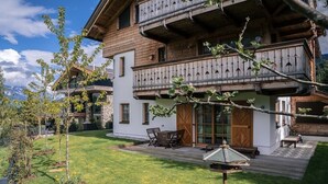 A two-story wooden house with a balcony, a patio with outdoor furniture, and a well-maintained lawn. - Steinberghaus Top 3 (Rain)