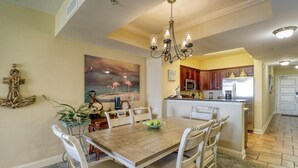 Un coin repas avec une table en bois et des chaises blanches, un lustre et un tableau représentant des flamants roses accroché au mur.