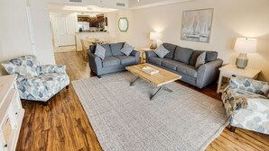 Sala de estar con un sofá gris, dos sillones, una mesa de centro y un tapete. Se ve una cocina al fondo.