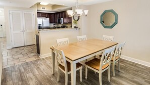 A dining area with a wooden table and white chairs, a kitchen with dark cabinets, and a decorative mirror on the wall. - Sip Coffee with a View: 3 Bedroom Oceanfront Condo (North Myrtle Beach)