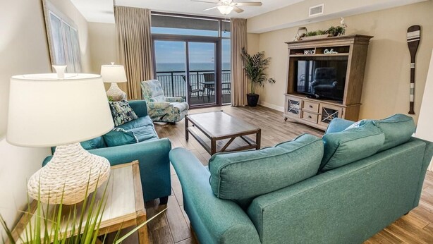 A living room with teal sofas, a wooden coffee table, a TV, and a large window overlooking the ocean. - Sip Coffee with a View: 3 Bedroom Oceanfront Condo (North Myrtle Beach)