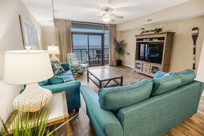 A living room with teal sofas, a wooden coffee table, a TV, and a large window overlooking the ocean.
