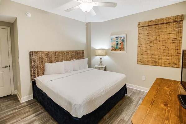 A bedroom with a bed, a nightstand, a lamp, a ceiling fan, a wooden table, and a wall-mounted artwork.
