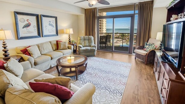 A living room with a sofa, armchairs, a coffee table, and a television. There are framed pictures on the wall, a ceiling fan, and a view of the beach through large windows. - Coastal Retreat 3 Bedroom Oceanfront 4th Floor (North Myrtle Beach)