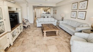 A spacious living room with a large sofa, a coffee table, and a TV on a cabinet. There is a ceiling fan, a kitchen in the background, and a dining area. - Relax in Style 3 Bdrm Condo with King and Queen (North Myrtle Beach)
