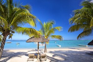 Ein Strand mit Palmen, einem Reetdachschirm und Liegestühlen.