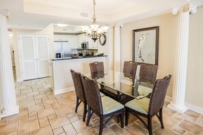 A dining area with a glass-top table and wicker chairs, a chandelier, and a kitchen in the background.