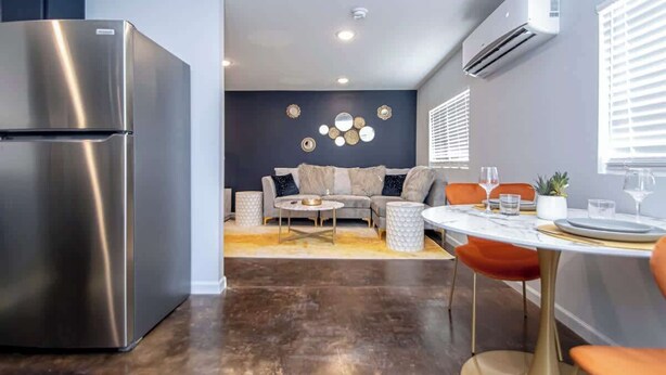 A modern kitchen with a stainless steel refrigerator, a dining area with a white table and orange chairs, and a living space with a sofa and decorative wall art. - Walk to Oaklawn Casino! Luxe Studio w/King Bed (Hot Springs)