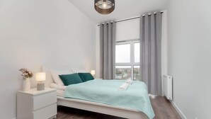 Ein modernes Schlafzimmer mit Bett, Nachttisch, Lampe und Blick auf die Stadt durch ein Fenster.