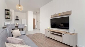 Ein modernes Wohnzimmer mit grauem Sofa, Holzboden, einem Fernseher auf einem Schrank und einer Pendelleuchte.