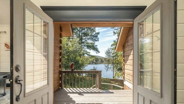 Puerta de madera abierta que da a una terraza con vistas a un lago y árboles.