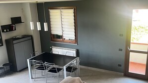 A room with a dining table and chairs, a sofa, a window with blinds, and a kitchenette area. - Modern Countryside Condo with Views (San Quirico In Collina)