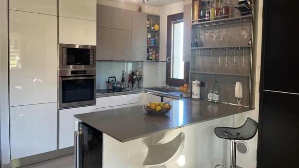 A modern kitchen with white cabinets, stainless steel appliances, a central island with bar stools, and a wine rack above the sink. - Modern Countryside Condo with Views (San Quirico In Collina)