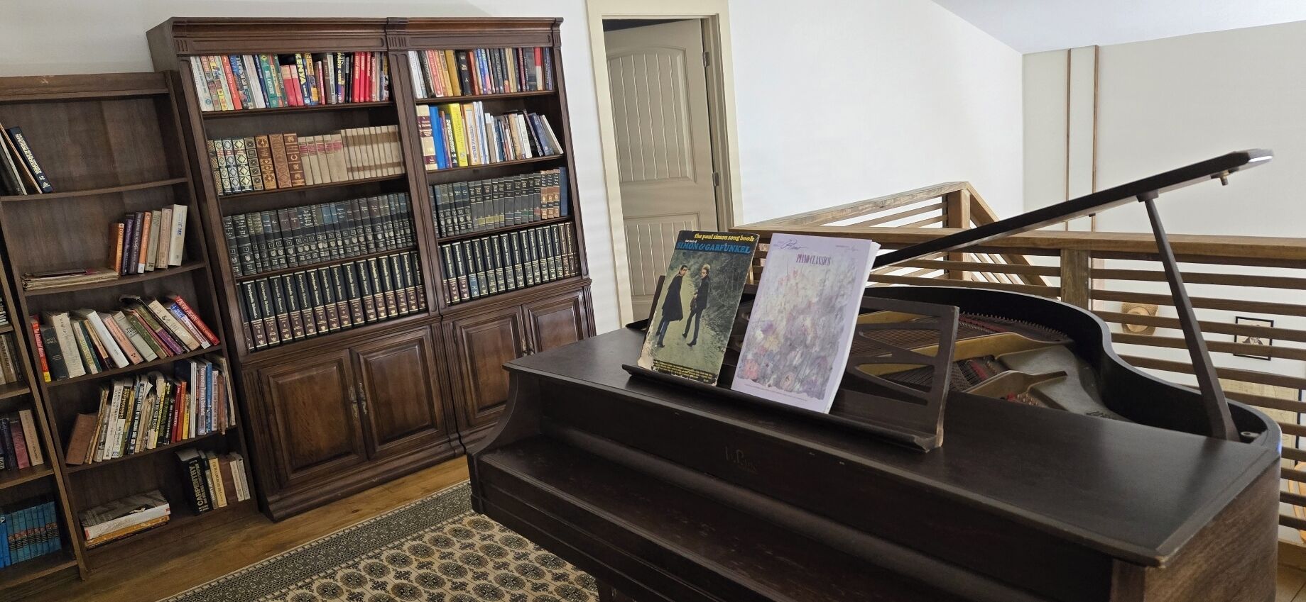 A piano with sheet music, a bookshelf filled with books, and a wooden staircase.