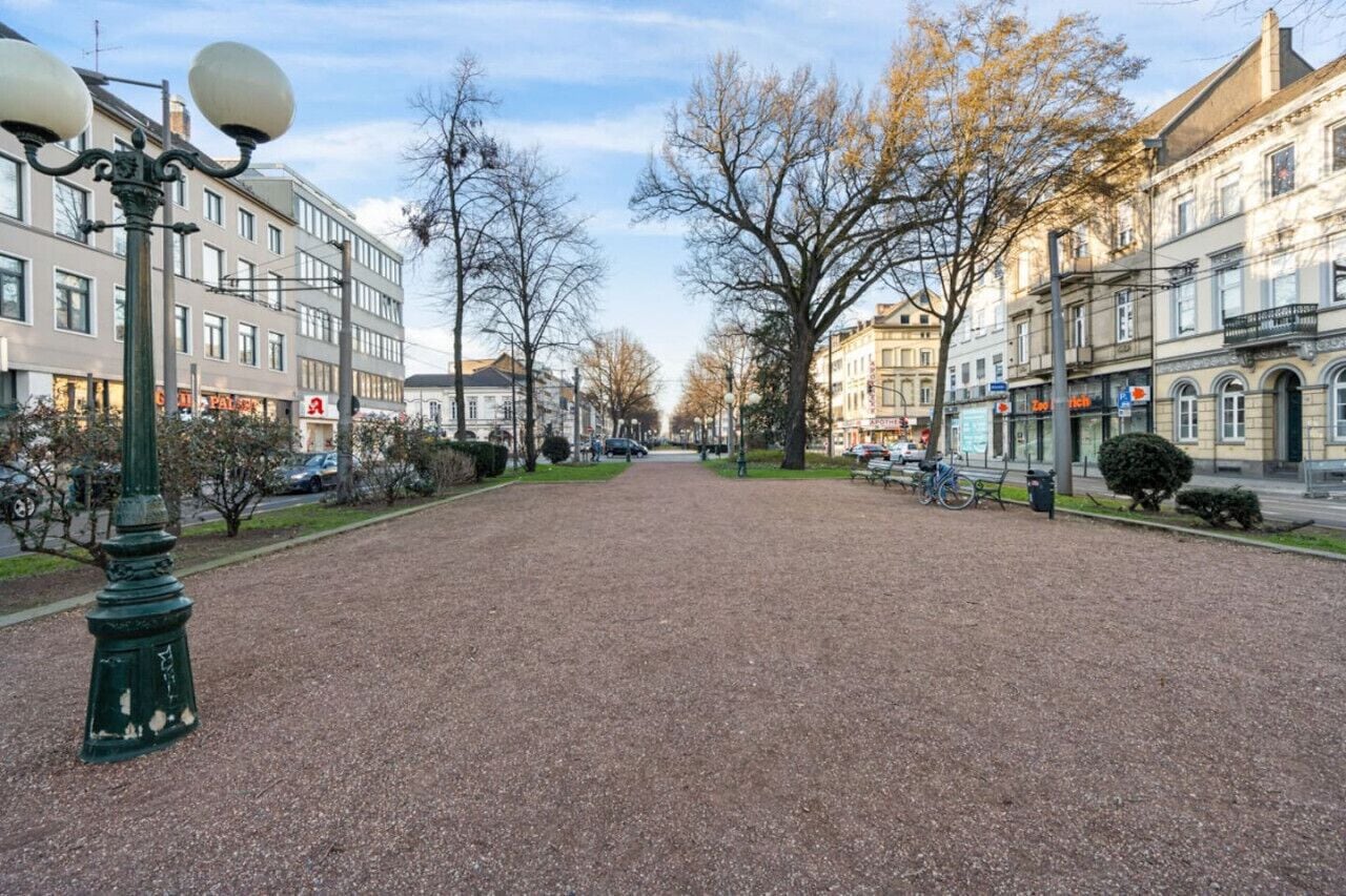 A paved pedestrian area with a lamp post, trees, and buildings in the background.