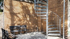 Pared exterior de madera con una escalera espiral de metal, un banco y una alfombra con estampado de cebra.