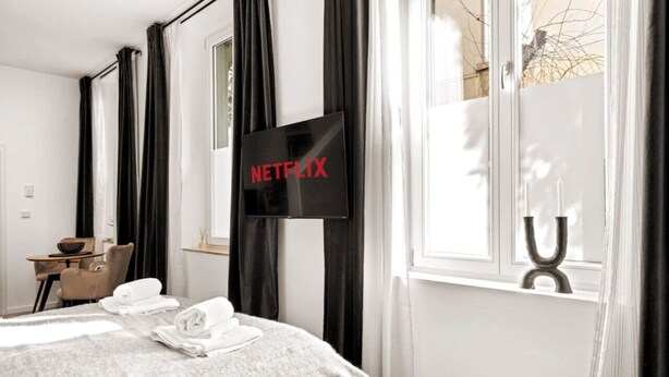 Una habitación de hotel con cama, televisor montado en la pared, ventana con cortinas y una mesita con un bol y una silla.