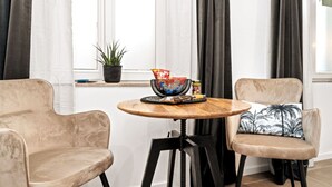 Una habitación con dos sillones beige, una mesita de madera con un bol y aperitivos, una planta en maceta y una ventana con cortinas.