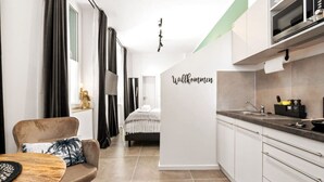 Una cocina moderna con armarios blancos, un microondas y un lavamanos. Se ve una cama al fondo con un cartel que dice "Willkommen".
