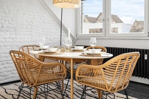Un coin repas avec une table ronde et quatre chaises en osier, un mur en briques blanches, une fenêtre donnant sur des immeubles et un radiateur noir.