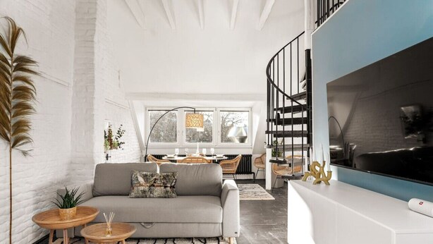 A modern living room with a grey sofa, wooden side tables, and a black staircase leading to a balcony.