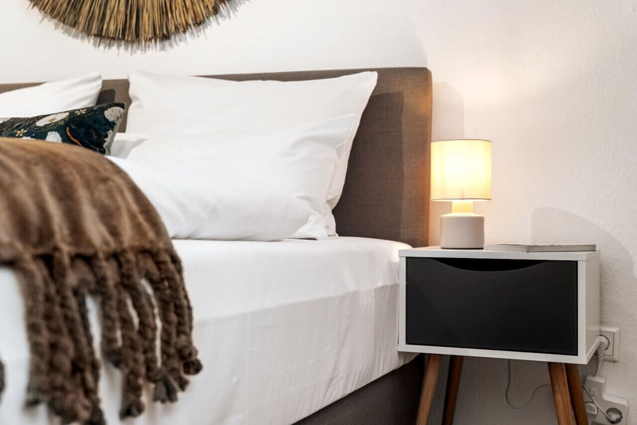 Una cama bien hecha con sábanas blancas y una manta marrón, una mesita de noche con una lámpara y un adorno en la pared sobre la cama.