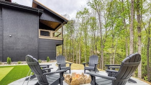 Een modern buitenzitgedeelte met Adirondack-stoelen en een vuurkuil, omringd door een verzorgd gazon en een bebost achtergrond.
