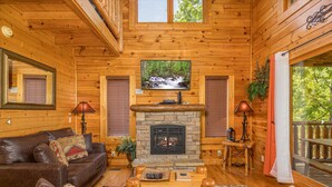 A cozy cabin interior with a stone fireplace, leather sofa, and a television mounted above the fireplace.