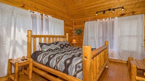 A wooden cabin bedroom with a bed, a nightstand, a lamp, and a window with curtains.