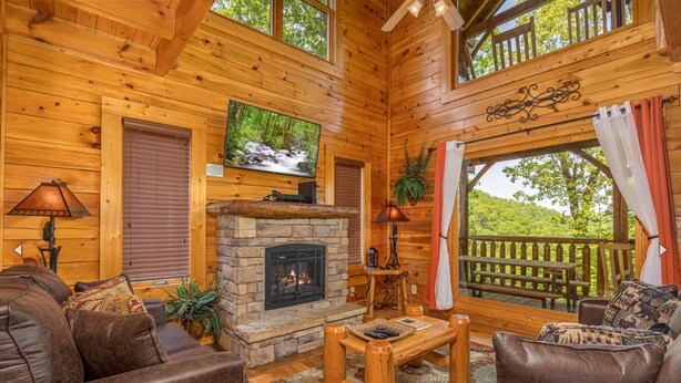 A cozy log cabin interior with a stone fireplace, leather sofas, a television, and a balcony with a wooden railing.
