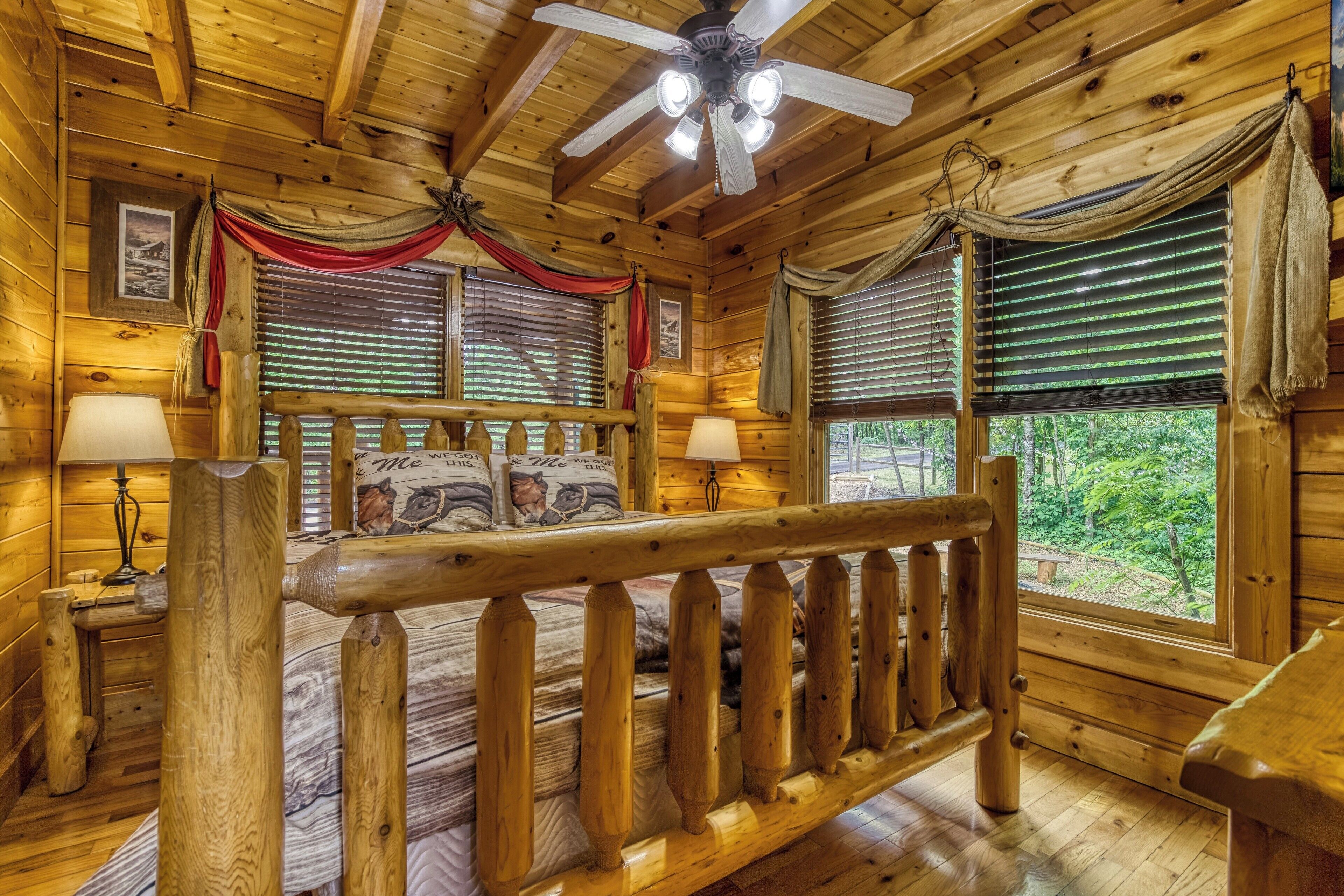 A wooden cabin bedroom with a bed, two bedside lamps, a ceiling fan, and windows with curtains.