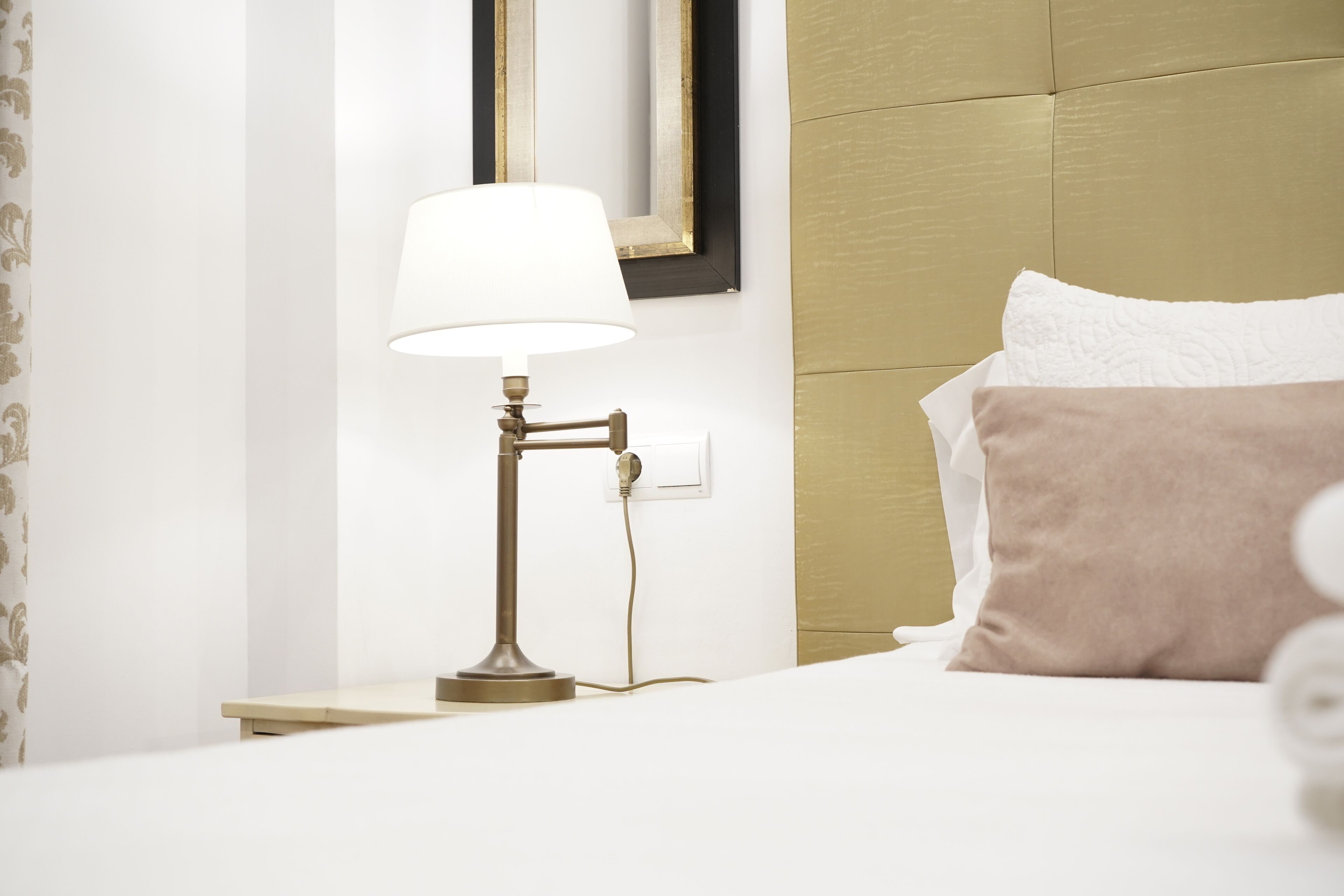 En pænt redt seng med hvid og beige pude, et natbord med lampe, og et spejl monteret på væggen.