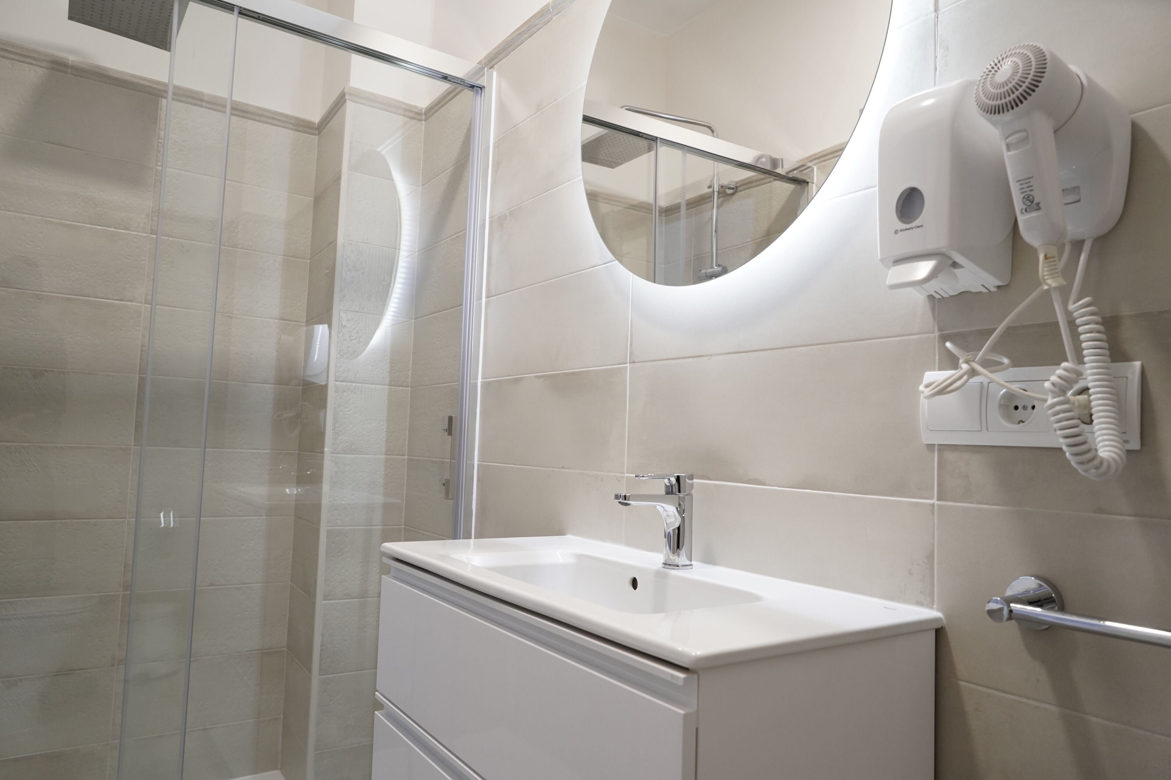 Une salle de bain moderne dotée d’une douche à parois de verre, d’un lavabo blanc avec un robinet chromé et d’un sèche-cheveux fixé au mur.