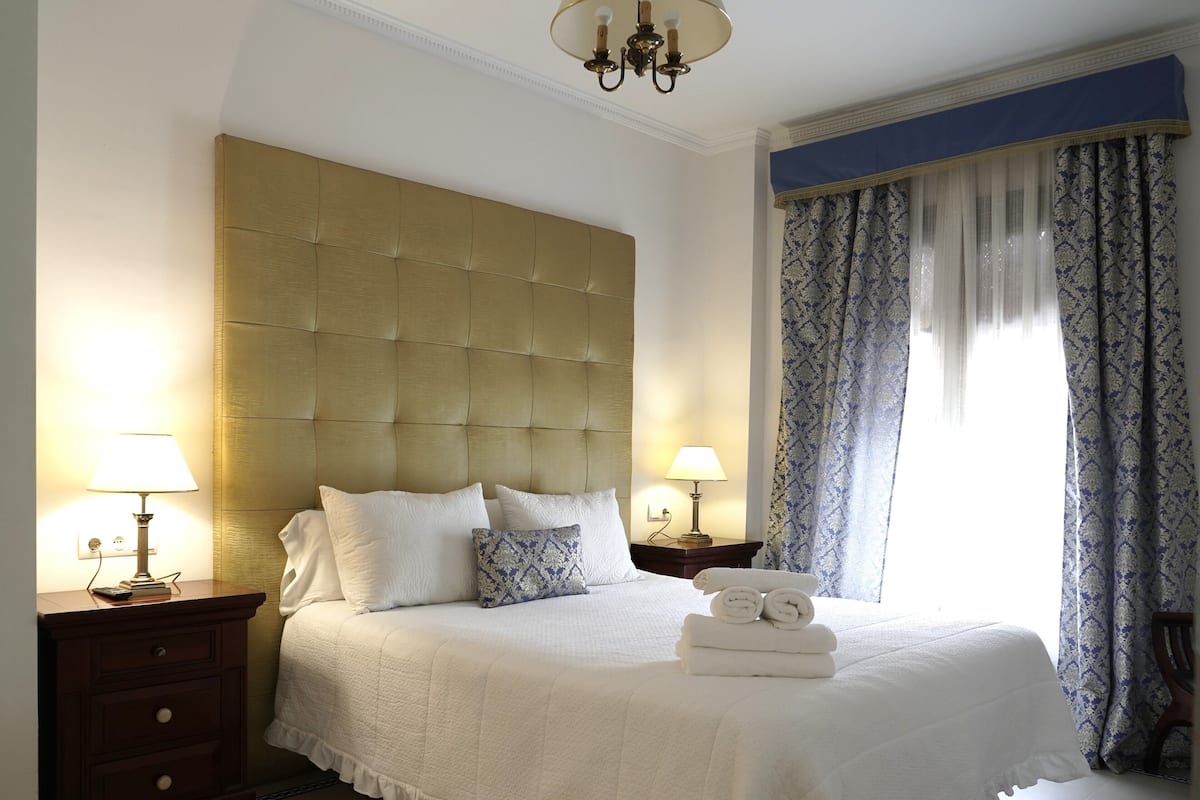 A neatly made bed with a tufted headboard, two bedside lamps, a wooden nightstand, and a window with patterned curtains.