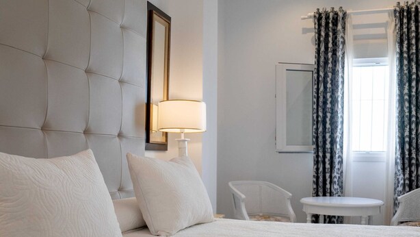 A hotel room with a large bed, a bedside table, a chair, a small table, and a window with curtains. - Palacete Real (Almonte)