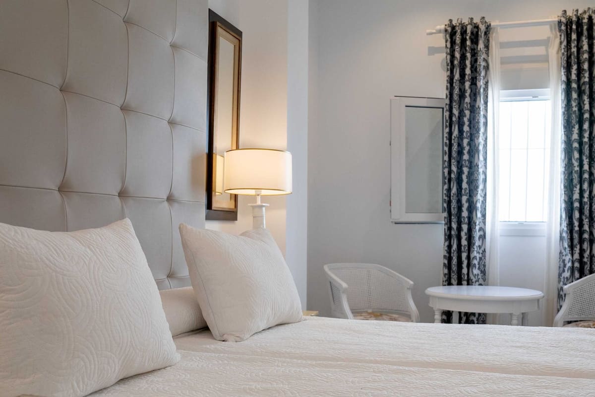 A hotel room with a large bed, a bedside table, a chair, a small table, and a window with curtains.