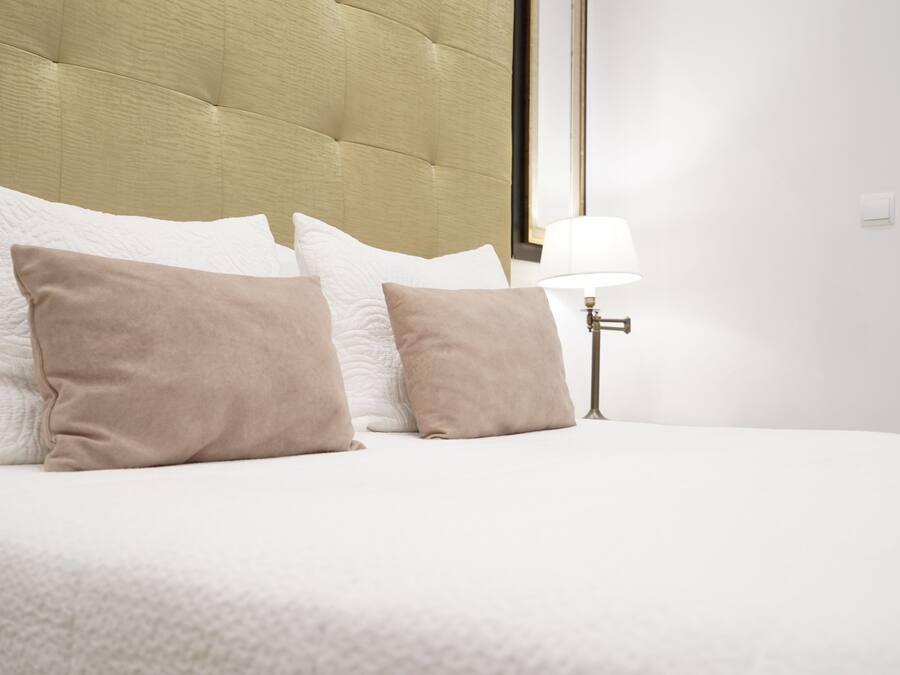 Una cama bien hecha con almohadas blancas y beige, un cabecero y una lámpara de noche.