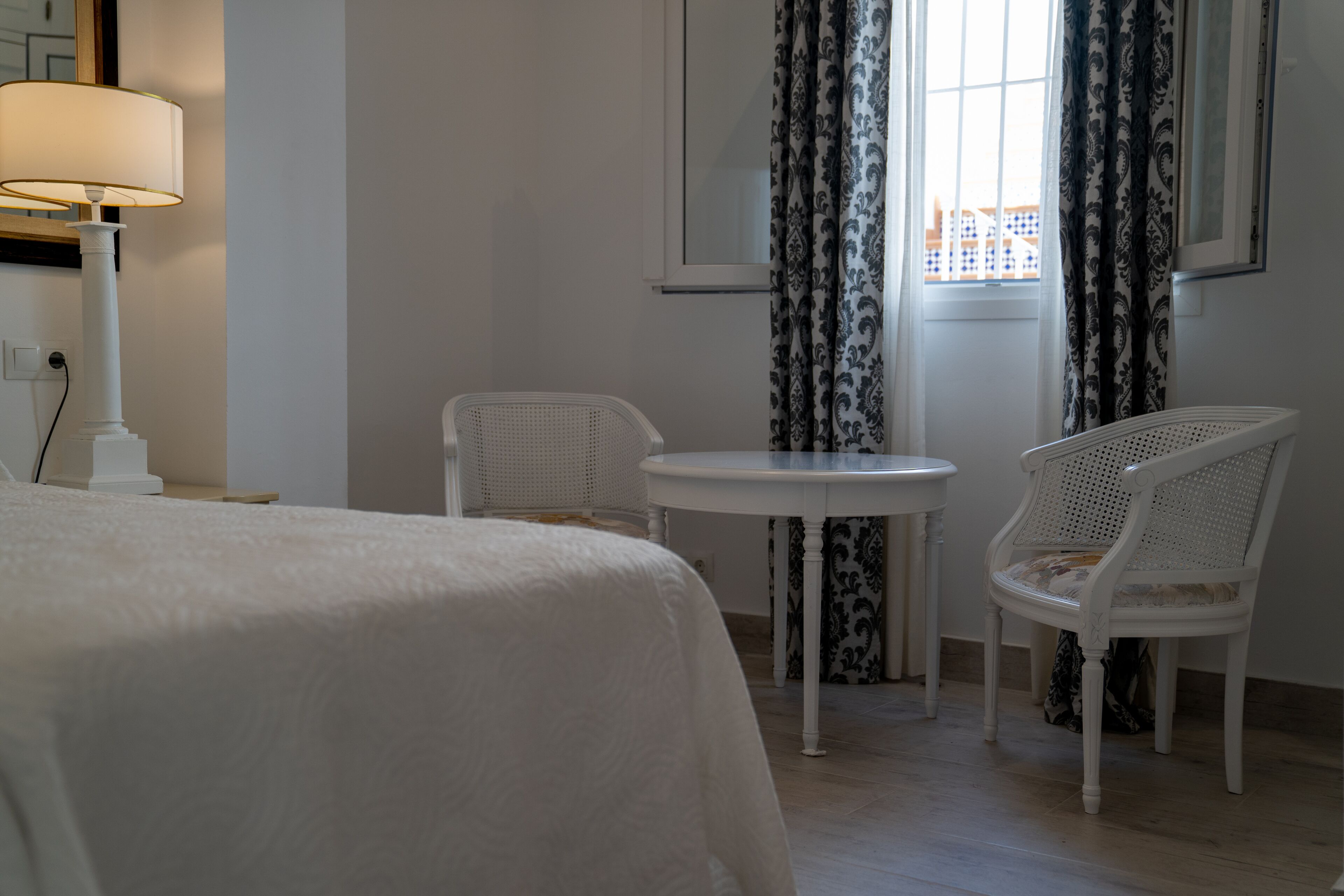 Et hotelværelse med en seng, et rundt bord, to fletstole, en lampe og et vindue med mønstrede gardiner.