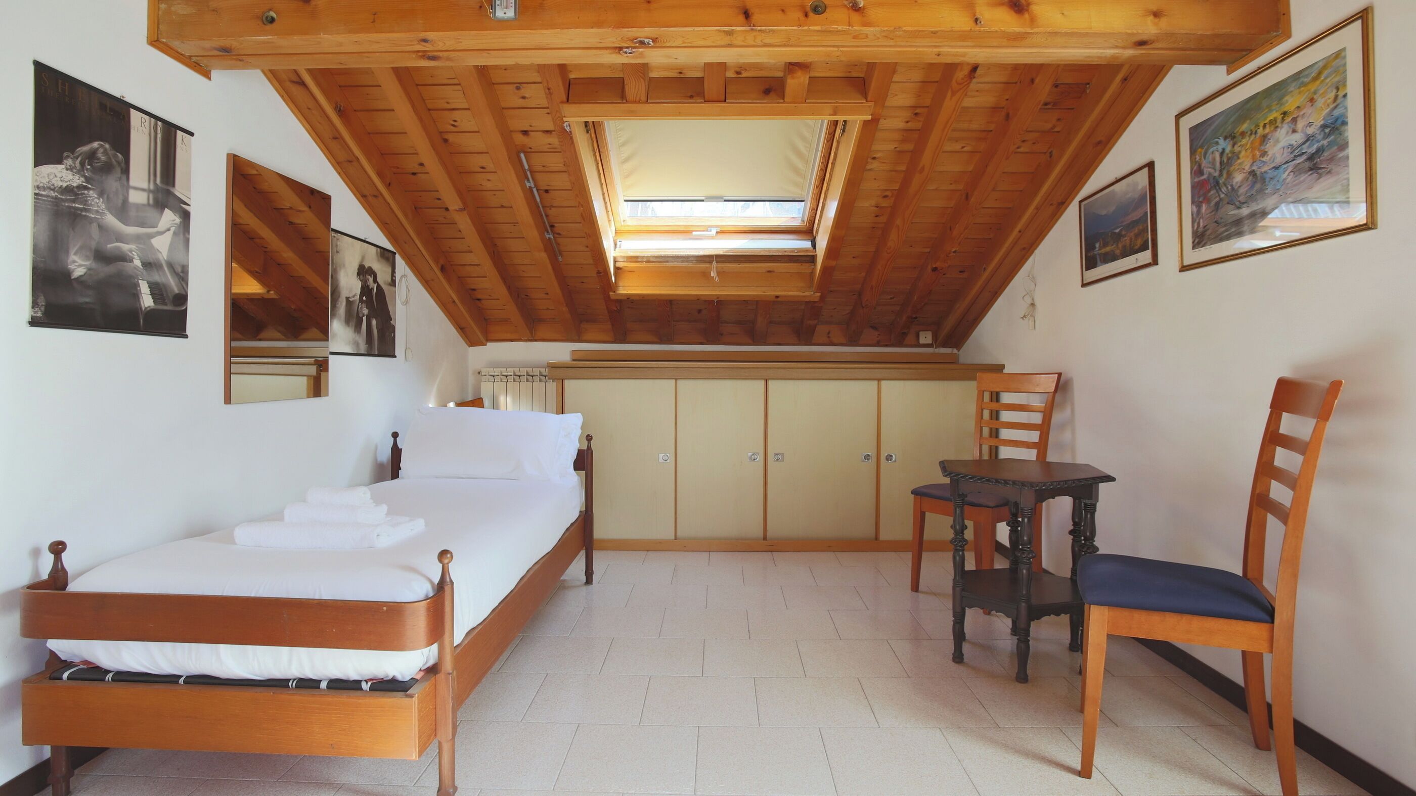 Quarto com cama, escrivaninha, cadeira e claraboia.
