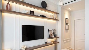Un salon moderne avec un téléviseur à écran plat fixé au mur, des étagères en bois ornées d’objets décoratifs et une porte blanche en arrière-plan.