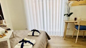 Une chambre d’hôtel avec un lit, une petite table sur laquelle se trouvent un vase et des verres, une chaise et une fenêtre ornée de rideaux légers.