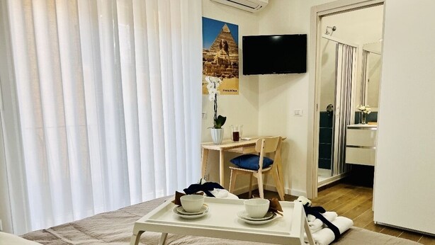 Une chambre d’hôtel avec un lit, une petite table sur laquelle se trouvent deux bols et un plateau, un bureau avec une chaise, un téléviseur fixé au mur et une salle de bain visible à travers une porte en verre.