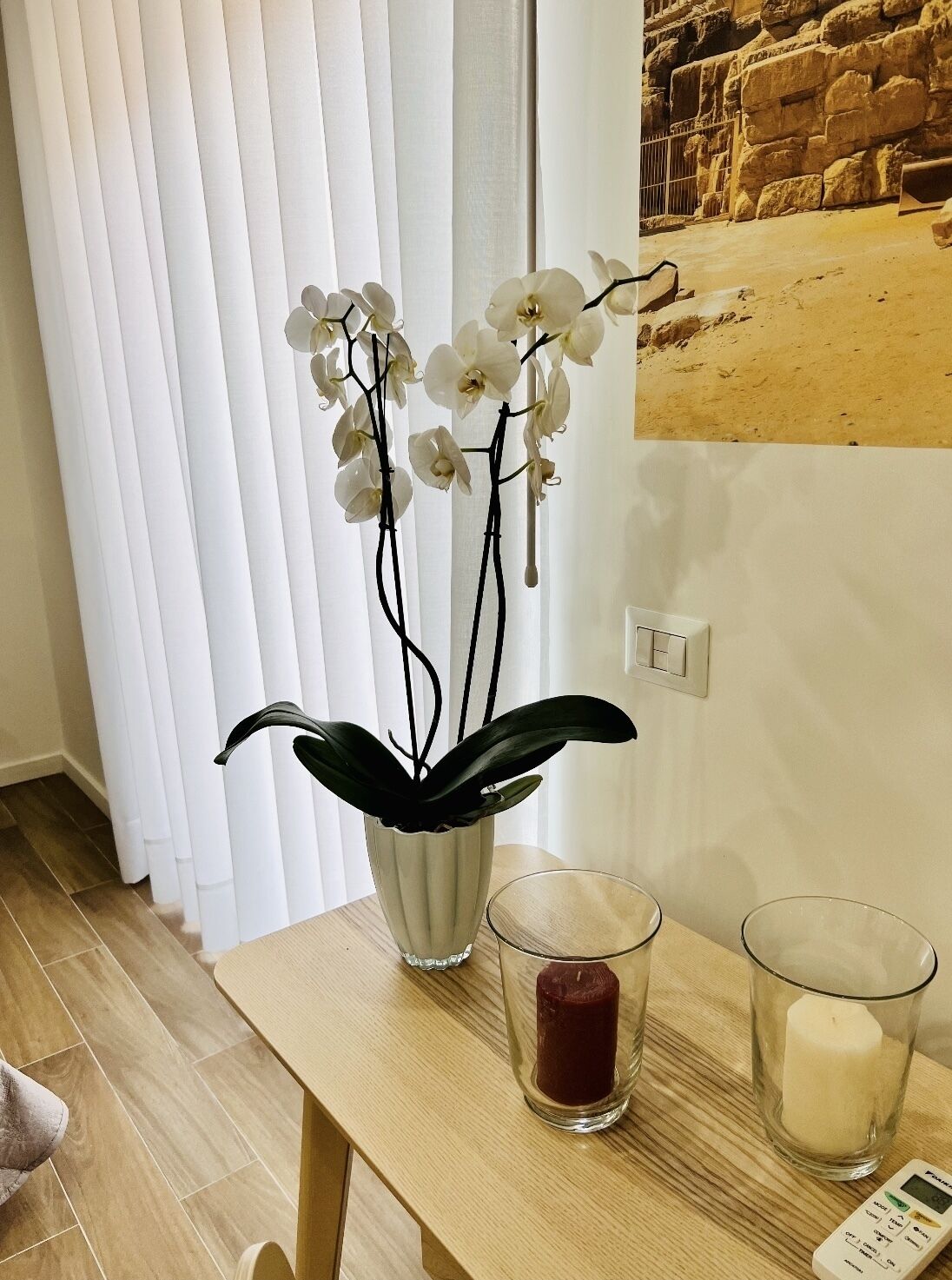 Una habitación con una mesa de madera, una planta de orquídea blanca en un florero blanco, dos velas y un termostato en la pared.