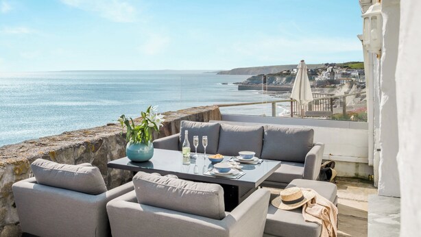 A seaside patio with a table set for dining, overlooking the ocean and a coastal town. - Loe Cottage (Helston)