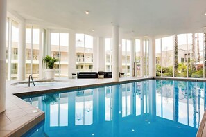 An indoor swimming pool with clear glass walls, a lounge area with seating, and a view of the surrounding buildings.