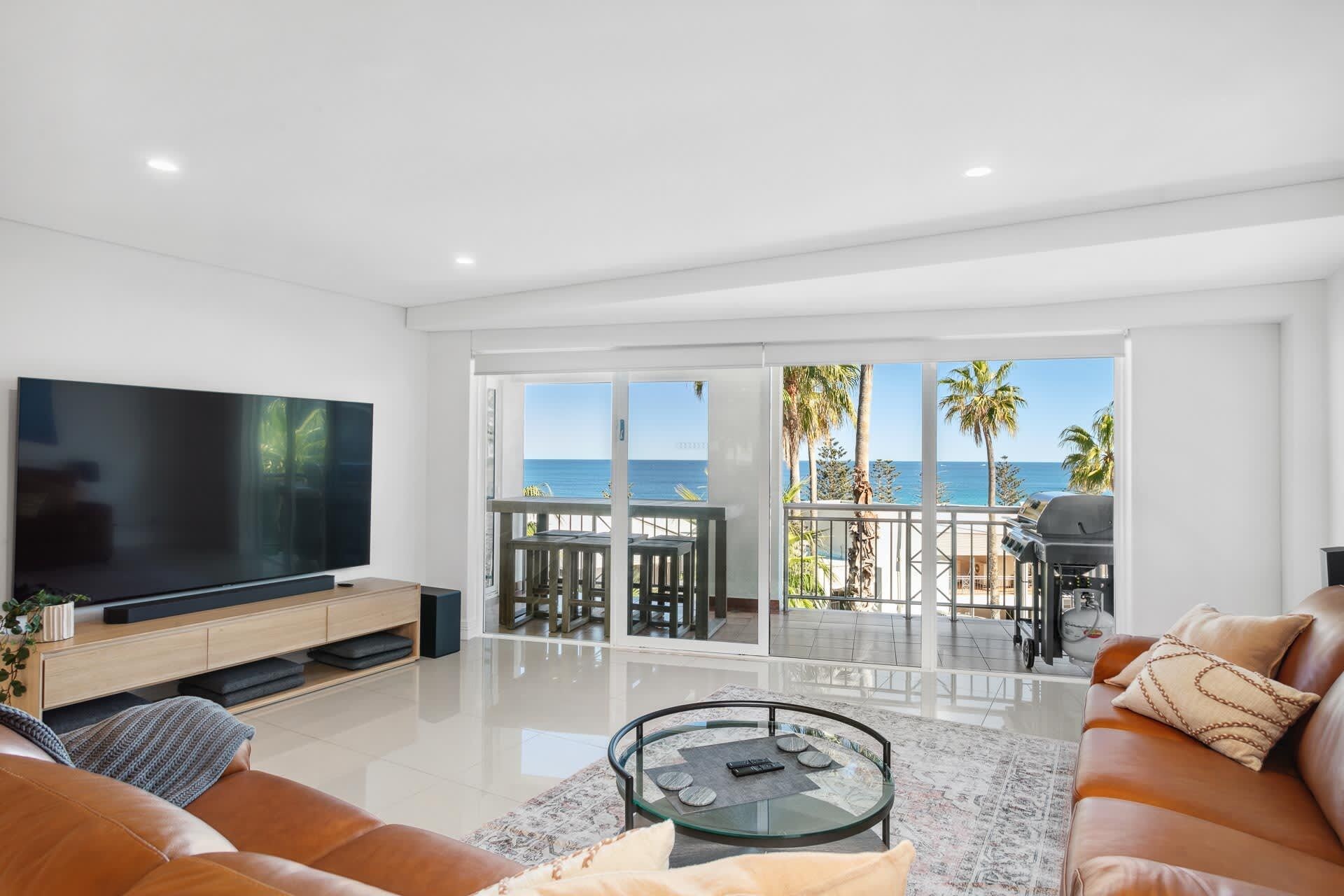 A modern living room with a large TV, leather sofa, and a glass coffee table. It features a balcony with a view of the ocean and palm trees.