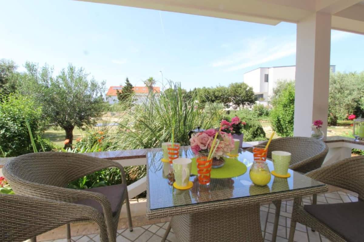Une table à plateau en verre, des chaises en osier, un vase rempli de fleurs et des boissons sur une terrasse offrant une vue sur la verdure et les bâtiments.