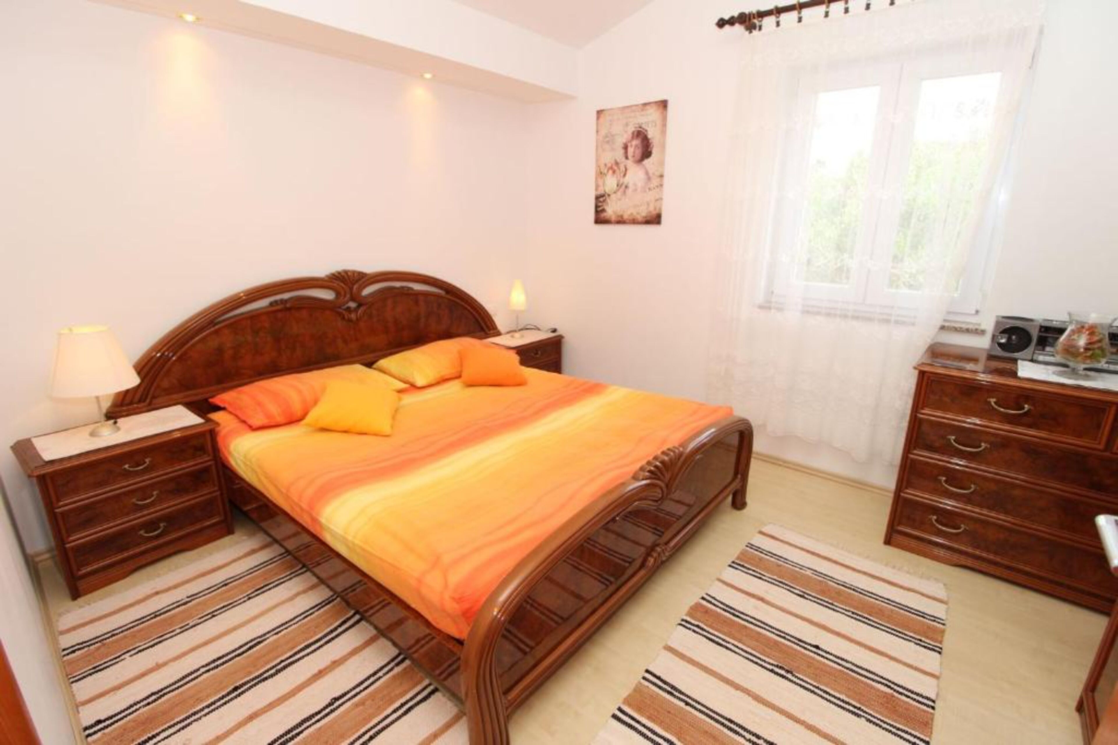 Un dormitorio con una cama de madera, alfombras a rayas, una mesita de noche, un aparador y una ventana con cortinas.