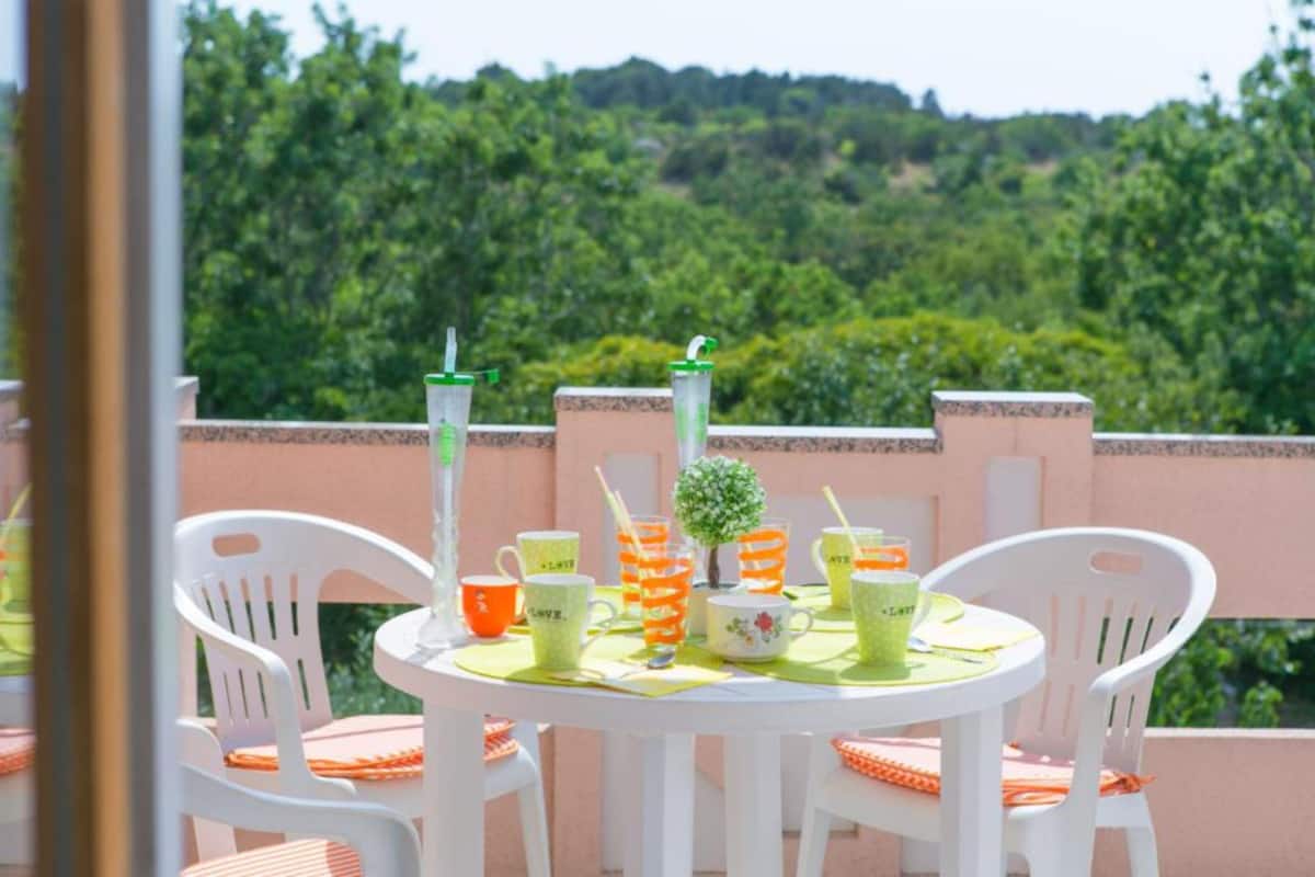 Une table ronde, entourée de chaises blanches et recouverte d’une nappe verte, est placée sur un balcon d’où l’on peut admirer un paysage verdoyant.