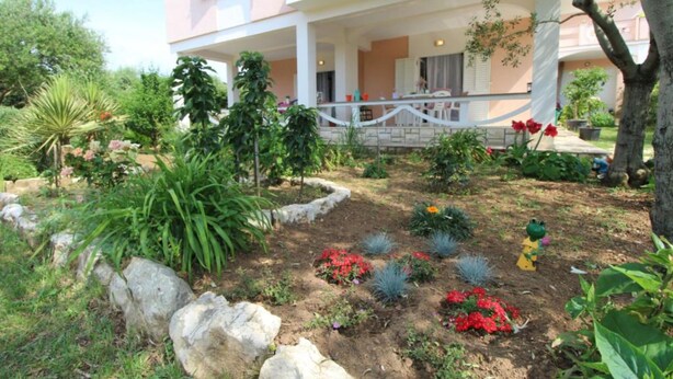 A pink building with a covered outdoor area, surrounded by a garden with various plants and flowers. - Apartments Danijela Novalja (Novalja)